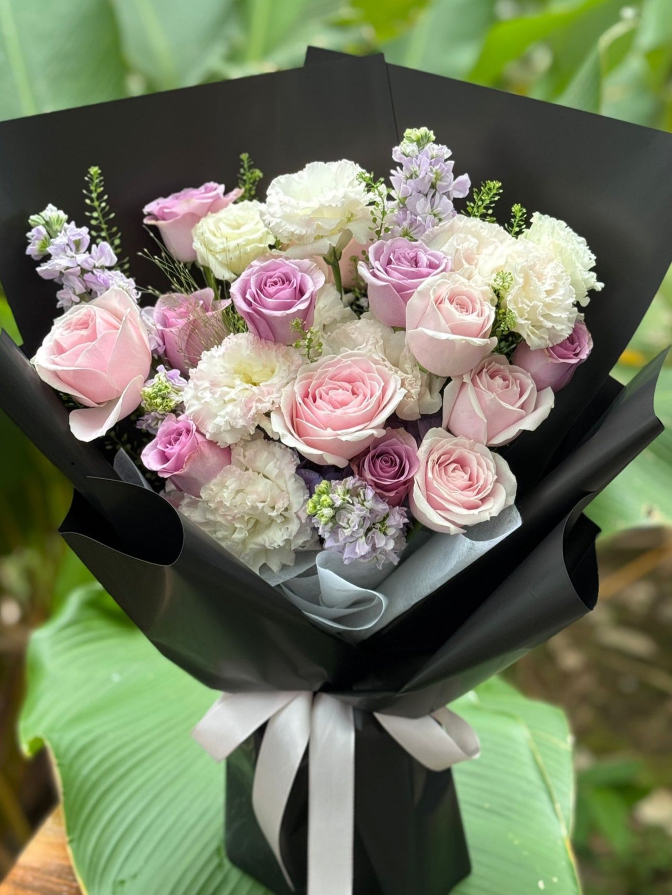 Bouquet of pink and white roses wrapped in black paper with a white ribbon, set against a green leafy background.